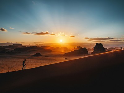 A person walking uphill on sand with the sunrise-sunset to his left.