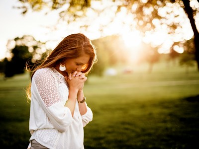 A girl with long hair praying outside with the sun shining in the background.