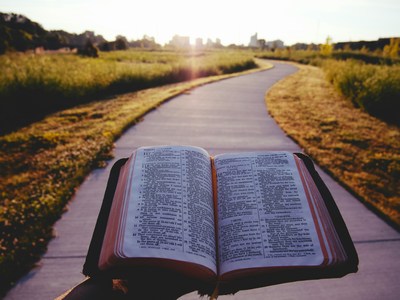 Someone holding an open Bible while walking on a sidewalk toward a city.